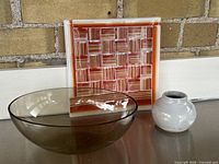 Smoked glass oval bowl alongside a square red and white patterned art glass plate and small light gray ceramic vase, displayed on metal surface against brick wall