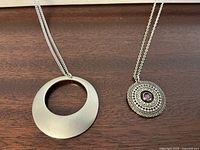 Two modern pewter pendant necklaces displayed on a wooden surface, showing the differing pendant designs.