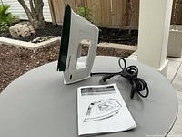 White and green steam iron standing upright on a round outdoor table with power cord and instruction manual beside it.