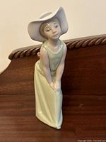 Front and angle view of Lladro porcelain figurine of girl in light green dress and white floppy hat, posed leaning forward.