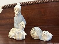 Two vintage Lladro porcelain figurines displayed on a dark wood surface: one girl with a lamb figurine and one duck with a basket figurine.