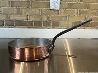 Side profile of the copper sauté pan showing copper exterior and black handle on a stainless steel surface.