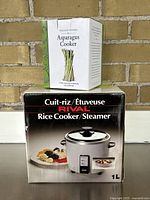 Williams-Sonoma Asparagus Cooker box stacked on top of Rival Rice Cooker/Steamer box against brick background.