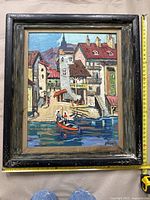 Front view of the framed oil painting showing a colorful town scene with water and a boat. The frame shows signs of wear and some paint loss visible on the canvas.