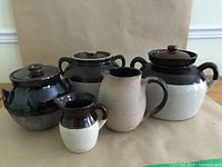 Five assorted stoneware items including 3 bean pots with lids and 2 pitchers, in brown and cream coloring, displayed on plain brown background.
