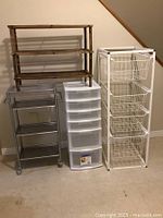 Four storage items including wooden shelf, metal rolling cart, plastic drawer unit, and wire basket unit lined up against wall on floor.