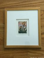 Full view of the framed artwork showing the floral print centered within double white mats and a light wood frame, placed on a wooden surface.