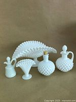Photo showing five white hobnail milk glass items including fan dish, vases and bottles grouped together on a flat surface with plain background.