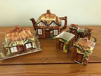 Photo showing the full lot on a wooden surface including teapot, cream and sugar set, covered butter, three smaller pieces, all cottage shaped with detailed hand painted earthy colors.