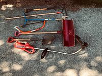Full view of the assorted yard tools laid on the ground including shovel, rake, cultivator, saw and electric trimmer.
