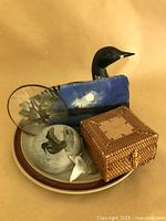 Photo showing grouped rustic decor items including loon decoy, woven box, painted slate, and plate, arranged on brown paper background.