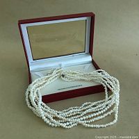 View of the pearl necklace resting on and beside its original rectangular jewelry box with transparent lid and white satin lining.