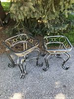 Pair of two differently sized metal patio tables with scrollwork galleries and curved legs, shown outdoors on pavement under a tree