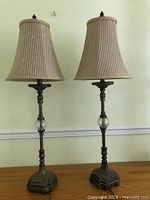 Full view of both tall skinny table lamps showing beige slub weave fabric shades, dark metal decorative bases with clear glass midsections, on a wooden surface against a plain wall.