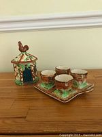 Four tiny egg cups styled as cottages on a matching tray with a covered jar topped by a rooster figurine, all hand painted in warm colors.