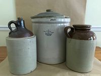 Front view of three stoneware pieces on a wooden table against neutral background showing the pickle jar with lid and two jugs with brown glazed tops and handles.