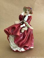 Side view of the Royal Doulton figurine showing the seated woman in flowing red dress, white ruffles, and red hat with blue accent.