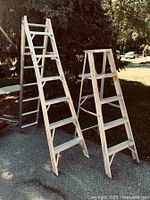 Two aluminum ladders standing outdoors, one tall multi-position extension ladder folded into A-frame next to a smaller 4-step ladder.
