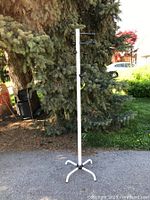 Full view of white tubular metal coat rack with three horizontal black ball-ended pegs and tripod base, photographed outdoors in daylight.