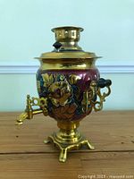 Front angled view of vintage enameled brass samovar showing colorful floral enamel decoration, brass spigot with black handle, and ornate feet.
