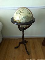 Full view of the terrestrial globe mounted on dark stained wooden tripod stand with three curved feet against a wall.