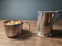 Two toddler cups side by side on a wooden surface with neutral background. The smaller cup is tarnished silver plated copper with handle and engraving, the taller is an English pewter cup with engraved text and decoration.