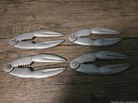 Four silver-colored metal lobster crackers arranged on a wooden surface, showing textured, serrated jaws and hinged handles.