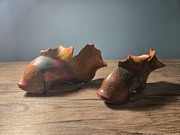 Two hand carved and hand painted wooden fish figurines displayed on a wooden surface against a gray wall, showing a side and front angle to highlight their detailed carving and paintwork.