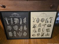 Two framed prints titled 'The Herb Garden' and 'The Vegetable Garden' with botanical illustrations on a wooden surface.