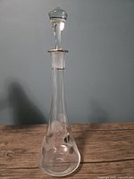 Clear crystal decanter with etched rose designs and silver leaf on both sides, standing on a wooden surface against a blue wall.