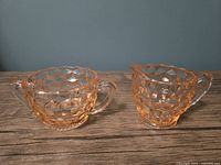 Two pink depression glass pieces, creamer on right and sugar bowl on left, showing cubist diamond cut pattern, scalloped edges, and pink hue on wooden surface with blue-gray background