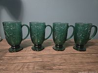 Four emerald green glass footed mugs arranged upright on wood surface, showcasing front embossed poinsettia patterns and handles.