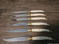 Top view of six Laguiole steak knives in a row, showing cream-colored handles with brass accents and stainless steel blades.