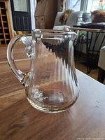 Front view of the clear blown glass mini pitcher showing rounded body and handle, placed on a wooden surface in a living room setting.