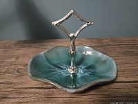 Blue Mountain Pottery snack dish with central silver-tone diamond-shaped handle, showing organic rounded shape and mottled blue-green glaze.