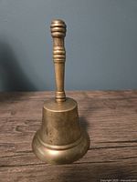 Full view of vintage solid brass bell with turned handle on wooden surface against gray wall.