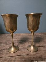 Side view of two vintage brass goblets with visible aged patina on a wooden surface against a blue wall.