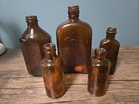 Group of five brown glass bottles of varying shapes and sizes on a wooden surface against a blue wall. Shows general size and design variety including largest bottle with embossed design.
