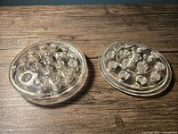 Two clear glass flower frogs on a wood surface, showing the top with multiple holes for flower arranging. One smaller with 16 holes and one larger with 19 holes, both in excellent condition.