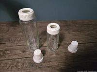 Photo shows two vintage Gerber glass baby bottles with white plastic caps on a wooden surface, and two additional white plastic caps placed in front of them.