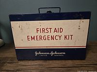 Closed exterior view of the vintage Johnson & Johnson metal first aid emergency kit, showing front text and handle.