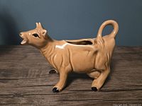 Vintage Kenmar ceramic cow creamer in brown with black details on hooves and eyes, cow's mouth open for pouring, with curved tail handle shown from side angle on a wooden surface.