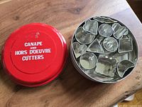 Open round red metal tin showing 12 metal canape/hors d'oeuvre cutters in various shapes.
