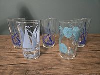 Five vintage juice glasses with different printed designs including blue sailboats, white sailboats, and blue flowers on clear glass.