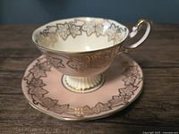 Front view of teacup with pink base and cream interior with gold leaf pattern and fluted footed base on saucer