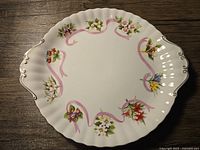 Top-down photo showing the hand-painted colorful provincial flowers and pink ribbon design on the bone china plate with scalloped edges and gold details on handles.