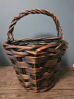 Front view of the dark brown and black woven wicker basket with twisted handle against a dark wall background.