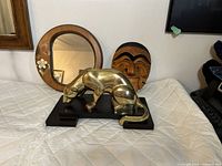 Image showing the brass panther sculpture in polished brass on black wood base, the 1970s round wood mirror with floral detail, and the vintage Korean wooden mask, all arranged on a white quilted surface.