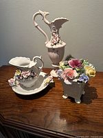 Photo showing the full set of three porcelain pieces: pink pitcher, white jug and saucer, and porcelain rose bouquet on wooden surface.