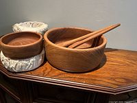 Teak salad bowls with two wooden serving utensils and nested smaller bowls wrapped in paper, placed on a wooden cabinet against a wall.